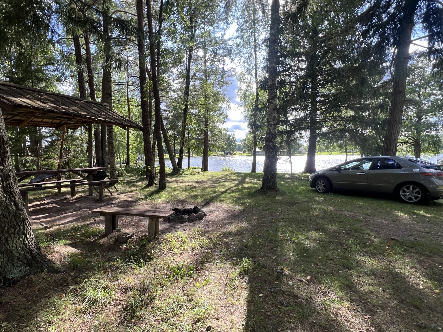 Shaded lakeside pitch with a picnic shelter, fire pit and car parked beside it