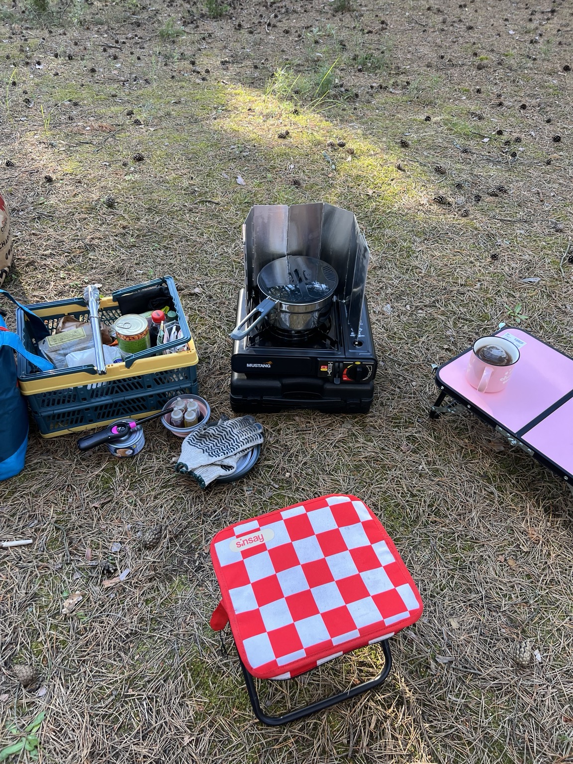 A compact campsite kitchen with a gas stove protected by a folding wind guard, crates and small tables