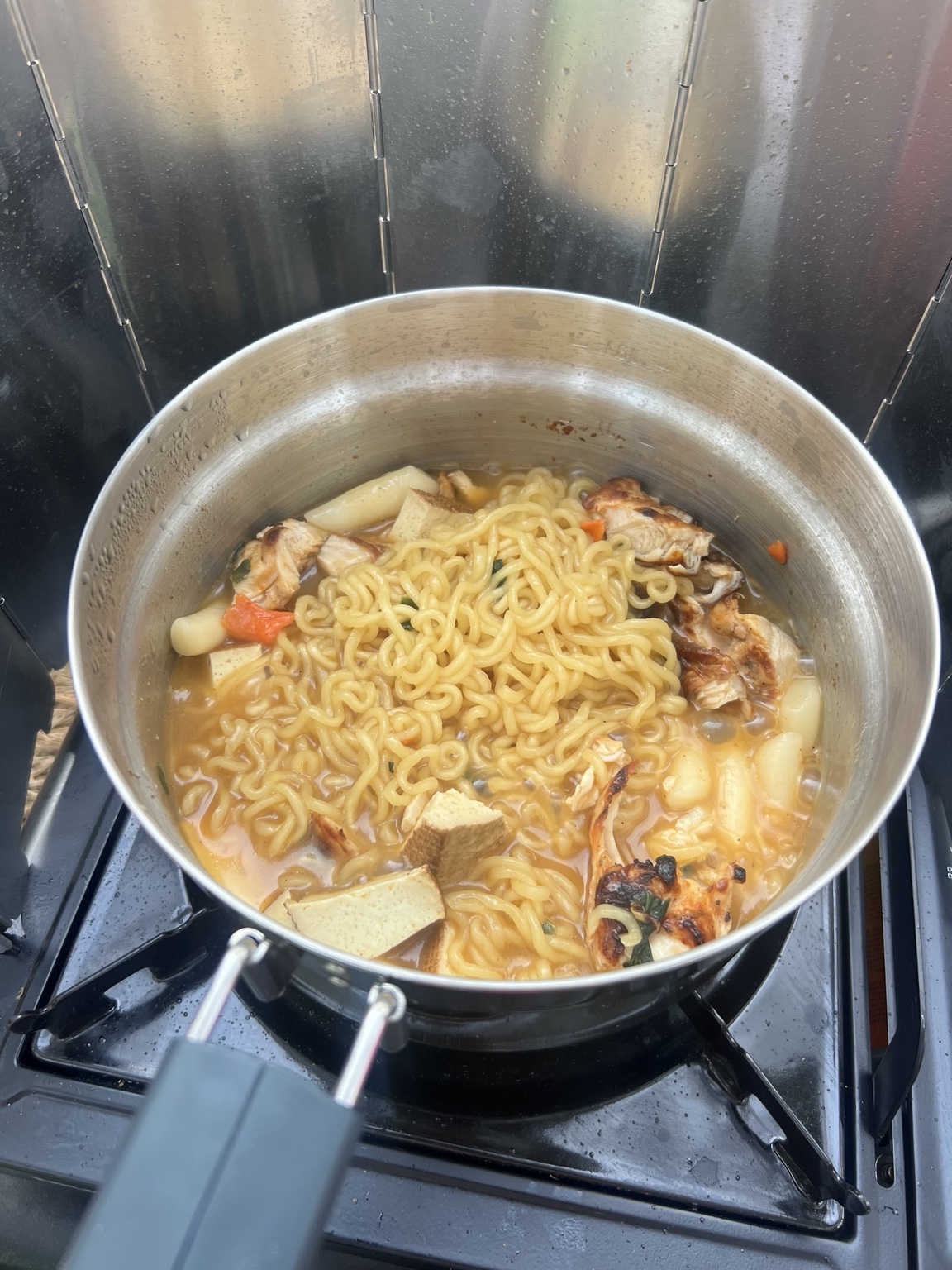 Ramen with leftover grilled chicken and tofu cooking on the campsite stove behind a wind guard